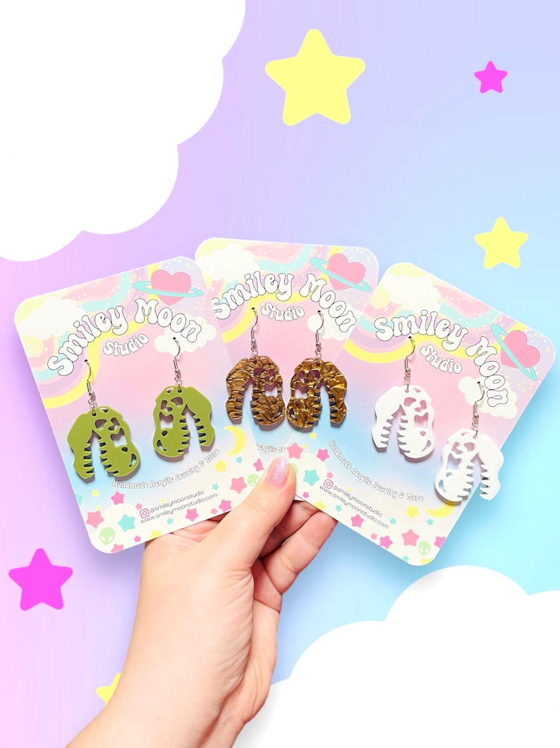 May include: Three pairs of acrylic earrings in green, gold, and white, each with a unique design. The earrings are displayed on colorful cards with the "Smiley Moon Studio" logo. The background features a pastel gradient with star and cloud accents.