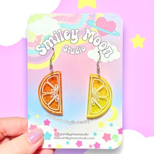 May include: A pair of orange and yellow glitter lemon slice earrings on a white card with the text "Smiley Moon Studio" and "Handmade Acrylic Jewelry & More".