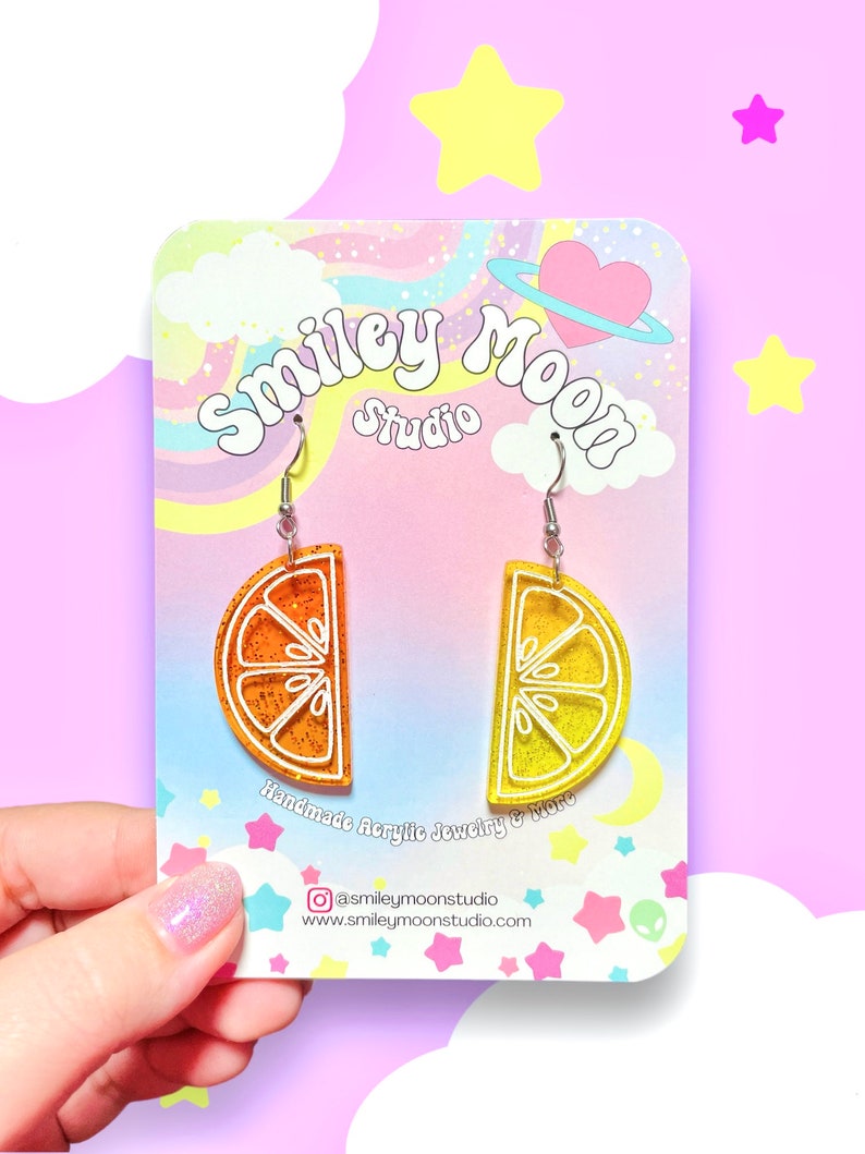May include: A pair of orange and yellow glitter lemon slice earrings on a white card with the text "Smiley Moon Studio" and "Handmade Acrylic Jewelry & More".