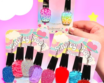 May include: A collection of colorful acrylic nail polish bottle earrings. The earrings feature various colors and patterns, including glitter, animal print, and solid hues. Each pair is attached to a card with the "Smiley Moon Studio" logo.