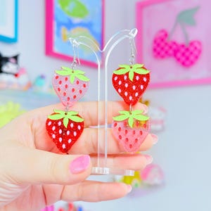 Peut inclure: Une paire de boucles d'oreilles pendantes compos&eacute;es de deux breloques fraises superpos&eacute;es. Les fraises en haut &agrave; gauche et en bas &agrave; droite sont rose clair paillet&eacute;es, tandis que celles en haut &agrave; droite et en bas &agrave; gauche sont rouge vif paillet&eacute;es.
