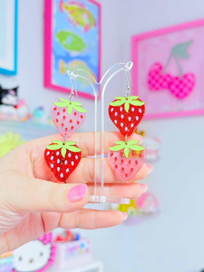 Peut inclure: Une paire de boucles d'oreilles pendantes compos&eacute;es de deux breloques fraises superpos&eacute;es. Les fraises en haut &agrave; gauche et en bas &agrave; droite sont rose clair paillet&eacute;es, tandis que celles en haut &agrave; droite et en bas &agrave; gauche sont rouge vif paillet&eacute;es.