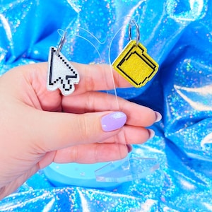 May include: A pair of novelty earrings, one shaped like a computer cursor and the other like a yellow file folder. The cursor earring is white with black pixelated details, while the file folder earring has a yellow glittery surface with black outlines.