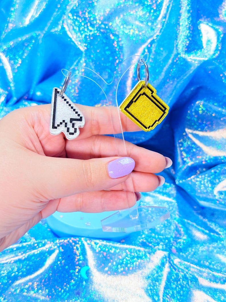 May include: A pair of novelty earrings, one shaped like a computer cursor and the other like a yellow file folder. The cursor earring is white with black pixelated details, while the file folder earring has a yellow glittery surface with black outlines.