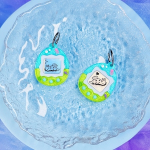 May include: Two egg-shaped charms featuring a digital pet interface design. Each charm has a bright blue and lime green border with a white screen displaying a black and white illustration of a sea slug. A silver metal jump ring is attached to the top.