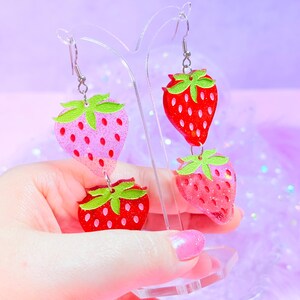 Peut inclure: Boucles d'oreilles pendantes compos&eacute;es de deux breloques fraises superpos&eacute;es. Les breloques sont en r&eacute;sine paillet&eacute;e dans des tons rouge et rose, avec des feuilles vertes sur le dessus. Elles sont suspendues &agrave; des crochets en m&eacute;tal argent&eacute;.
