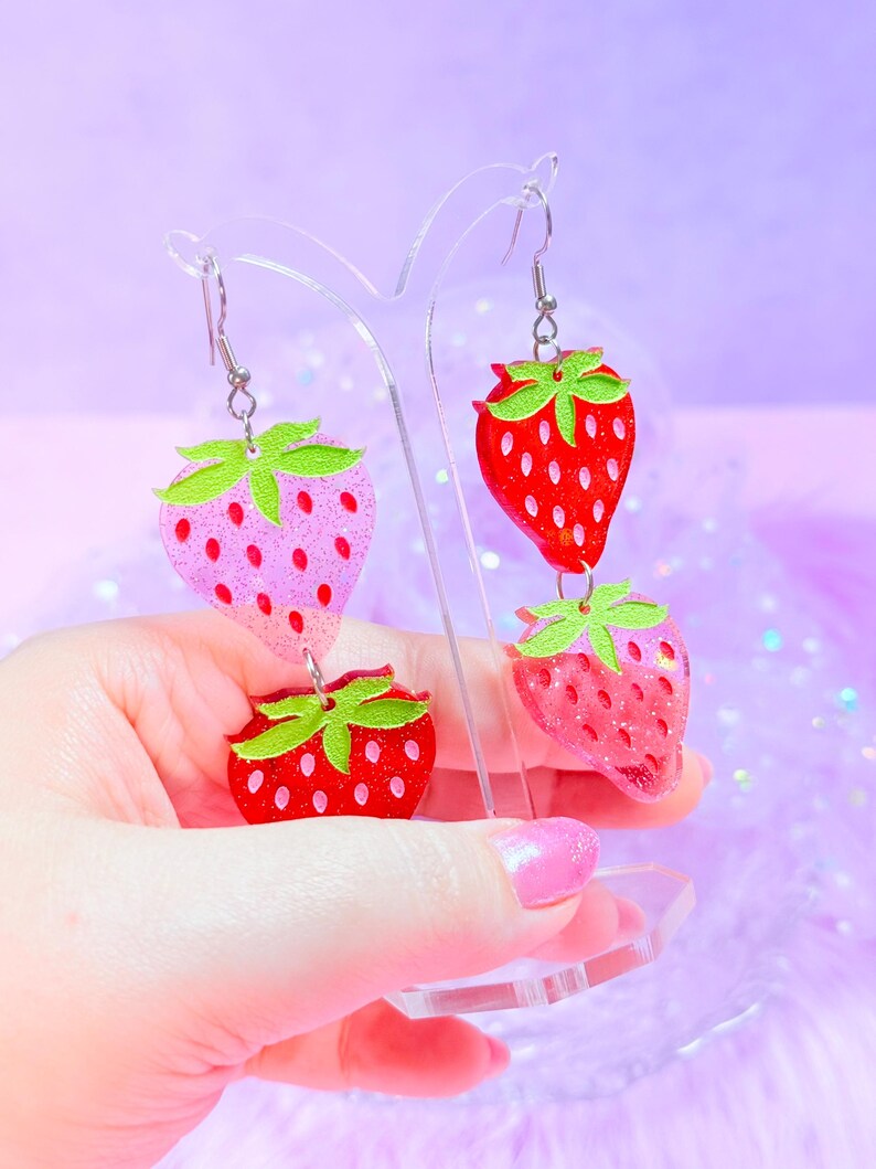 Peut inclure: Boucles d'oreilles pendantes compos&eacute;es de deux breloques fraises superpos&eacute;es. Les breloques sont en r&eacute;sine paillet&eacute;e dans des tons rouge et rose, avec des feuilles vertes sur le dessus. Elles sont suspendues &agrave; des crochets en m&eacute;tal argent&eacute;.