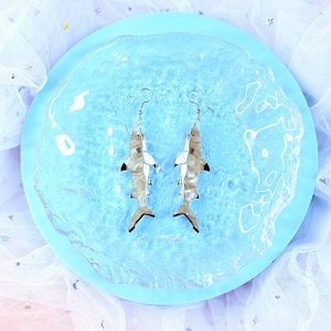 May include: Shark-shaped earrings with silver hooks. The earrings have a white belly, a grey and white body, and black fins. They are presented on a light blue, round plate, against a backdrop of sheer, pastel fabric.