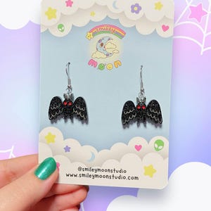 K&ouml;nnte beinhalten: Ein Paar schwarze Fledermaus-Ohrringe mit roten Augen und Glitzer, die an silbernen Haken h&auml;ngen. Die Ohrringe werden auf einer hellblauen Karte mit einem Cartoon-Mond- und Wolken-Design pr&auml;sentiert. Die Karte enth&auml;lt auch den Text "smiley moon" und Social-Media-Informationen.
