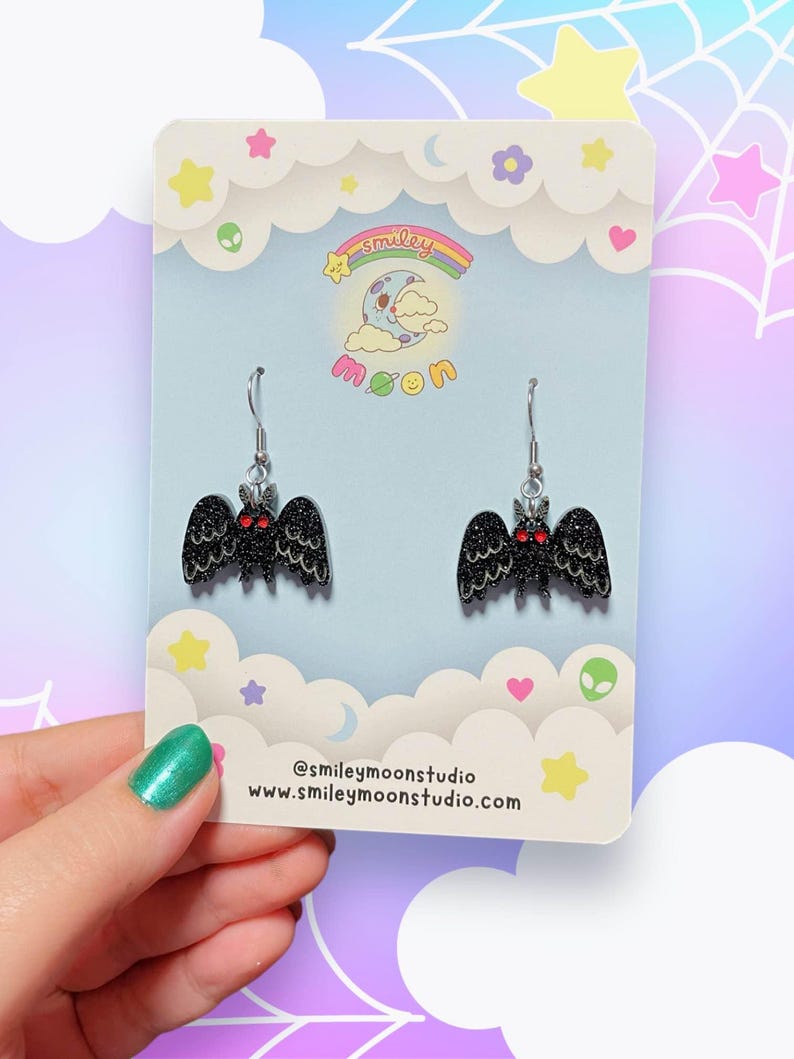 K&ouml;nnte beinhalten: Ein Paar schwarze Fledermaus-Ohrringe mit roten Augen und Glitzer, die an silbernen Haken h&auml;ngen. Die Ohrringe werden auf einer hellblauen Karte mit einem Cartoon-Mond- und Wolken-Design pr&auml;sentiert. Die Karte enth&auml;lt auch den Text "smiley moon" und Social-Media-Informationen.