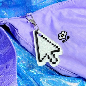 May include: A silver-colored cursor-shaped keychain with black pixelated details hangs from a silver clasp. The keychain is attached to a lavender bag with a small flower graphic. The background features a shiny, blue fabric.