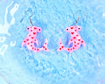 May include: A pair of dangle earrings featuring light pink hammerhead shark charms. Each shark charm is decorated with small red heart patterns and has a black eye. The earrings are attached to silver-toned metal fishhook wires for pierced ears.