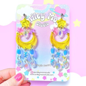 May include: Yellow crescent moon earrings with iridescent blue and pink charms, and a clear crystal charm. The earrings are on a white card with the text "Smiley Moon Studio" and a social media handle.