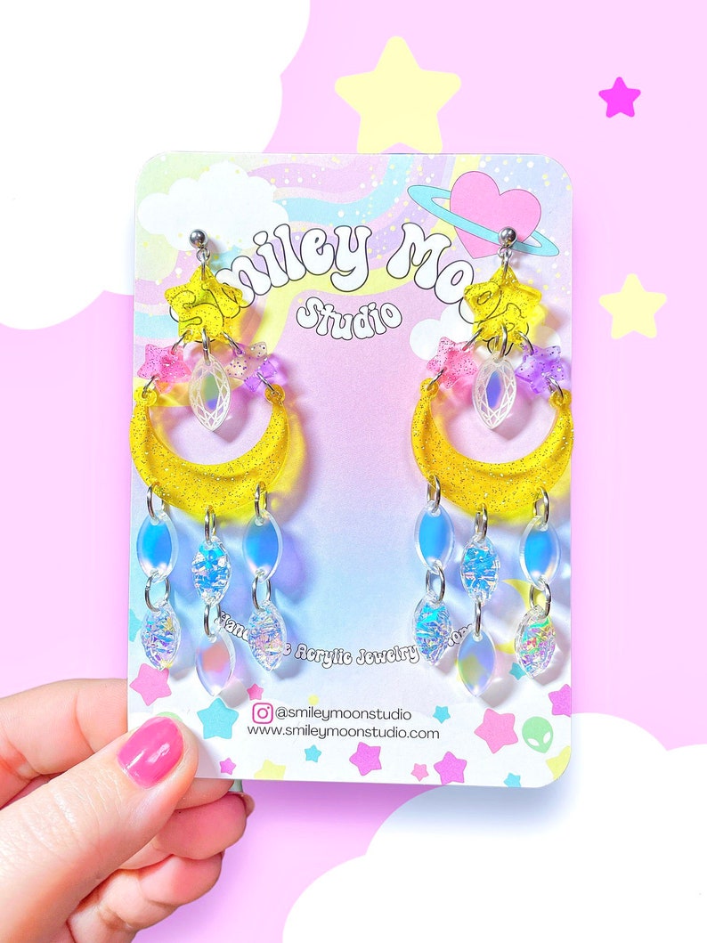May include: Yellow crescent moon earrings with iridescent blue and pink charms, and a clear crystal charm. The earrings are on a white card with the text "Smiley Moon Studio" and a social media handle.