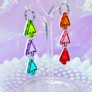 May include: Dangle earrings featuring a vertical stack of three pixelated computer cursor charms each. The left earring has green, purple, and teal cursors, while the right earring has orange, pink, and red cursors. Each charm is made of glittery acrylic with a black outline and hangs from a silver-toned metal hoop.