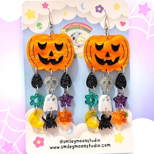 May include: A pair of orange and black Halloween-themed dangle earrings featuring a pumpkin, spider, ghost, star, and flower charms. The earrings are on a white card with the text "@smileymoonstudio www.smileymoonstudio.com".