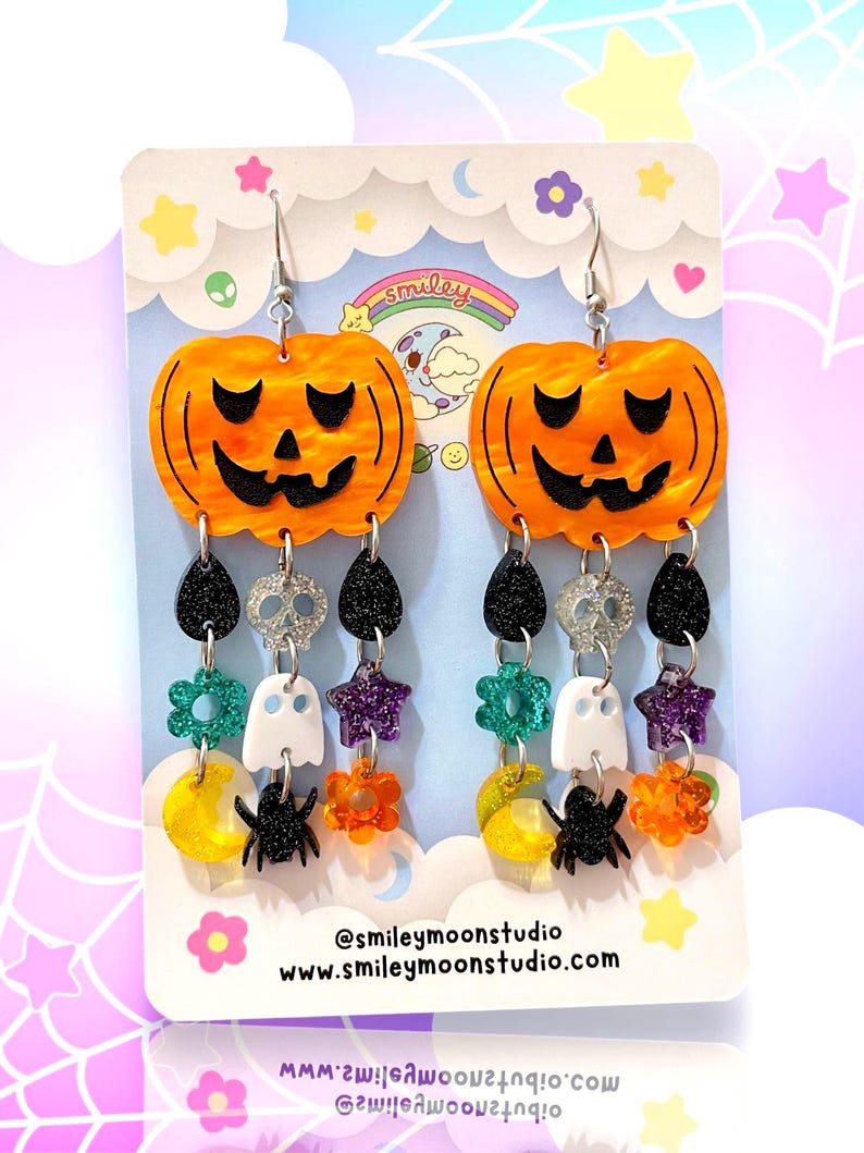 May include: A pair of orange and black Halloween-themed dangle earrings featuring a pumpkin, spider, ghost, star, and flower charms. The earrings are on a white card with the text "@smileymoonstudio www.smileymoonstudio.com".