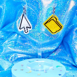 May include: A pair of pixelated earrings. One earring is a white and black computer cursor, the other is a yellow file folder. The earrings hang from silver hoops. They are displayed on a clear stand with a light blue base decorated with star and moon-shaped confetti.
