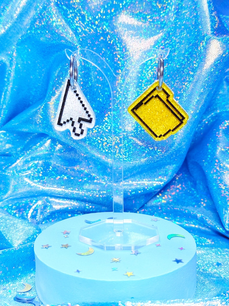 May include: A pair of pixelated earrings. One earring is a white and black computer cursor, the other is a yellow file folder. The earrings hang from silver hoops. They are displayed on a clear stand with a light blue base decorated with star and moon-shaped confetti.