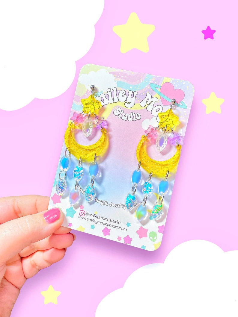 May include: A pair of yellow glitter crescent moon earrings with iridescent blue and pink accents. The earrings are dangling with iridescent blue teardrop-shaped charms. The earrings are displayed on a white card with the text "Smiley Moon Studio" and a social media handle.