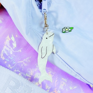 May include: A white beluga whale charm with black outline hangs from a silver clasp attached to a light blue zip. The zip is on a light blue jacket. A small embroidered flower is visible.