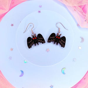 K&ouml;nnte beinhalten: Ein Paar schwarze Mottenohrringe mit Glitzerakzenten und roten Augen. Die Ohrringe haben ein detailliertes Fl&uuml;geldesign und sind an silberfarbenen Ohrhaken befestigt. Die Ohrringe werden auf einer hellblauen Oberfl&auml;che mit dekorativen Sternen und Monden pr&auml;sentiert.