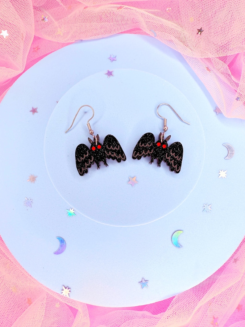 K&ouml;nnte beinhalten: Ein Paar schwarze Mottenohrringe mit Glitzerakzenten und roten Augen. Die Ohrringe haben ein detailliertes Fl&uuml;geldesign und sind an silberfarbenen Ohrhaken befestigt. Die Ohrringe werden auf einer hellblauen Oberfl&auml;che mit dekorativen Sternen und Monden pr&auml;sentiert.