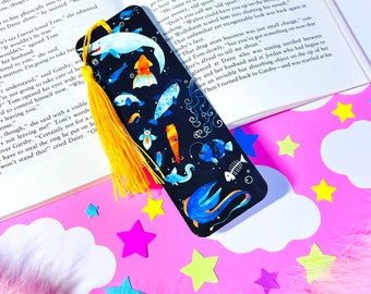 May include: A black bookmark with a yellow tassel features a colorful illustration of various sea creatures, including a shark, squid, fish, and a seahorse. The bookmark is surrounded by white clouds and pink stars.