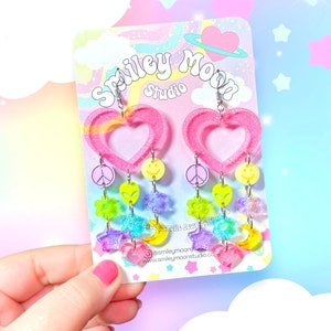 May include: Pink heart-shaped dangle earrings with rainbow-coloured charms, including peace signs, smiley faces, stars, moons, and alien heads. The earrings are on a white card with the text "Smiley Moon Studio" and a website address.