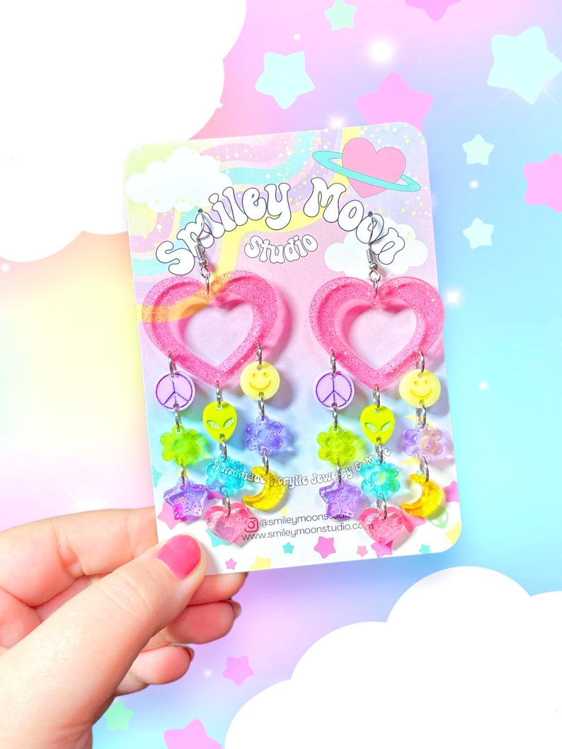 May include: Pink heart-shaped dangle earrings with rainbow-coloured charms, including peace signs, smiley faces, stars, moons, and alien heads. The earrings are on a white card with the text "Smiley Moon Studio" and a website address.
