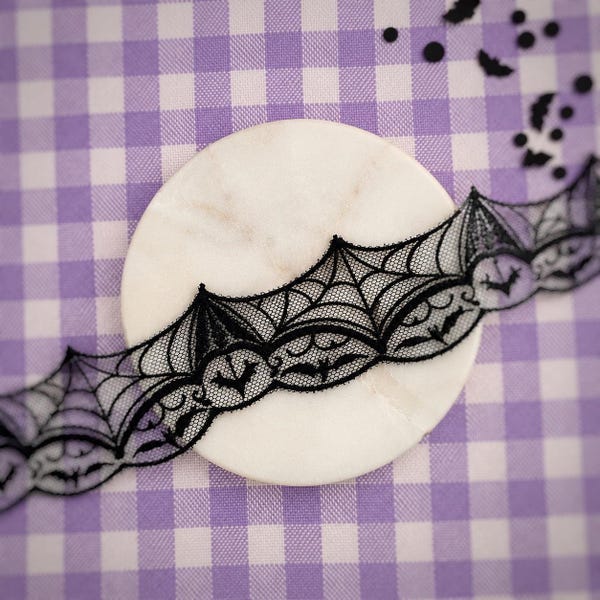 May include: A strip of black lace trim featuring a repeating pattern of spider webs and small bat silhouettes. The lace has a scalloped edge and is positioned across a smooth, round, white marble disc. The background is a light purple gingham fabric.