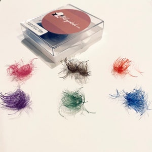 Könnte beinhalten: Sechs kleine Haufen bunter Wimpernextensions auf einer weißen Oberfläche. Die Extensions sind in den Farben Pink, Braun, Grün, Blau, Lila und Orange. Die Extensions sind in einem Kreis um eine kleine weiße Box mit dem Text "5D 0.07CC 8-15mm" auf der Seite angeordnet.