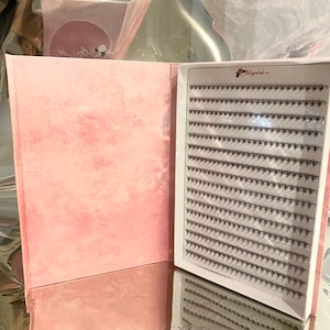 May include: A pink, open box displaying a grid of individual black eyelash extensions. The box has a pink, textured cover and a white interior. The brand name "Belladash" is visible on the upper right corner of the eyelash grid.