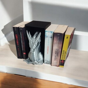 Könnte beinhalten: Silberne Drachen-Buchstützen, die einen Stapel Bücher halten. Der Drache liest ein Buch. Die Bücher haben Titel in Deutsch und Englisch.