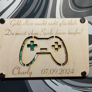 Könnte beinhalten: Eine Holzkarte mit einem Laser-geschnittenen Design eines Videospiel-Controllers. Die Karte hat den Text "Geld allein macht nicht glücklich. Du musst schon Spiele davon kaufen!" und "Charly 07.09.2024".
