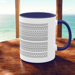 Könnte beinhalten: Weiße Keramiktasse mit marineblauem Innenraum und Griff. Die Tasse hat ein sich wiederholendes schwarz-weißes geometrisches Muster auf der Außenseite. Die Tasse steht auf einer Holzoberfläche mit Blick auf das Meer im Hintergrund.