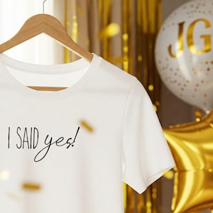 以下が含まれることがあります： 白いTシャツに、黒い文字で「I SAID yes!」と書いてあります。Tシャツは木製ハンガーにかけられています。背景はぼやけていて、金色と白い風船が見えます。