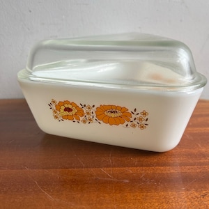 May include: A vintage white casserole dish with a clear glass lid. The dish has a floral design in orange and yellow. The dish is likely made of glass and is a classic kitchenware item.