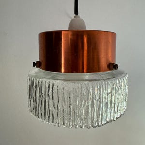 May include: A vintage pendant light with a copper-coloured metal shade and a textured glass bottom. The light hangs from a black cord and a white ceiling fixture. The glass has a unique, vertical, ridged pattern.