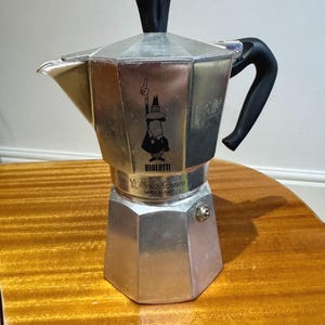 May include: A silver-coloured stovetop espresso maker with a black handle and top. The coffee pot has an octagonal base and a cartoon man logo. The words "Bialetti" and "Moka Express" are visible. Made in Italy.