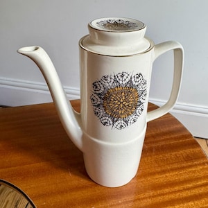 May include: Cream ceramic coffee pot with a black and gold floral design. The pot has a long spout, a handle, and a lid. The design is centered on the pot and lid, with gold trim.