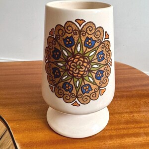 May include: Cream ceramic vase with a pedestal base. The vase has a symmetrical floral design in brown, orange, blue, and green. The design is centered on the vase, adding a decorative element. The vase is suitable for displaying flowers.