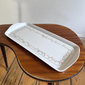 May include: A rectangular white ceramic serving tray with rounded corners and raised edges. The centre of the tray features a delicate decorative border pattern consisting of thin, intertwined pink ribbons and small green vines with tiny pink flowers.