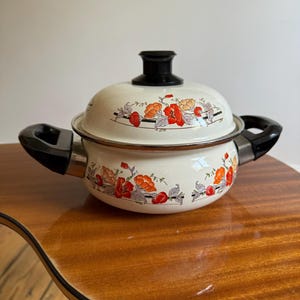 May include: Cream-coloured enamel pot with a matching lid. The pot and lid are decorated with a floral pattern in red and orange. The pot has black handles and a black knob on the lid. A vintage kitchenware item.