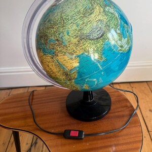 May include: A spherical world globe featuring a detailed map with blue oceans and yellow and green landmasses. The globe is supported by a clear plastic meridian arc and a black circular base. A black power cord with a red switch is attached.