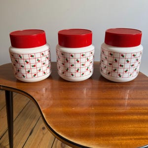 May include: Three vintage white canisters with red lids. The canisters feature a repeating pattern of red and green cherries on a white background. The canisters are on a wooden table.
