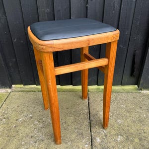 May include: A vintage wooden stool with a square, black padded seat. The stool has four slender, tapered legs and a horizontal support bar. The wood has a warm, natural finish, and the stool is set against a dark background.