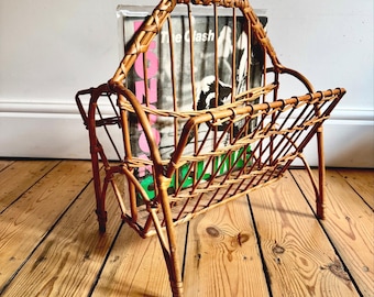 May include: A vintage wicker magazine rack with a handle, holding a vinyl record. The rack is made of woven brown wicker and has a rectangular shape with a slatted design. The record sleeve has the text "The Clash".