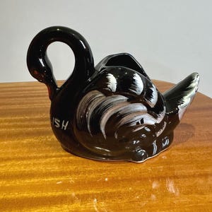 May include: A black ceramic swan-shaped creamer with white and grey feather detailing. The creamer has a curved handle and a small opening for pouring. The word "ISH" is printed on the side.