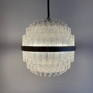 May include: A spherical pendant light fitting featuring a clear, textured glass shade with vertical rod-like protrusions. A dark metal band encircles the centre of the globe, which is suspended from a single black cord against a plain background.